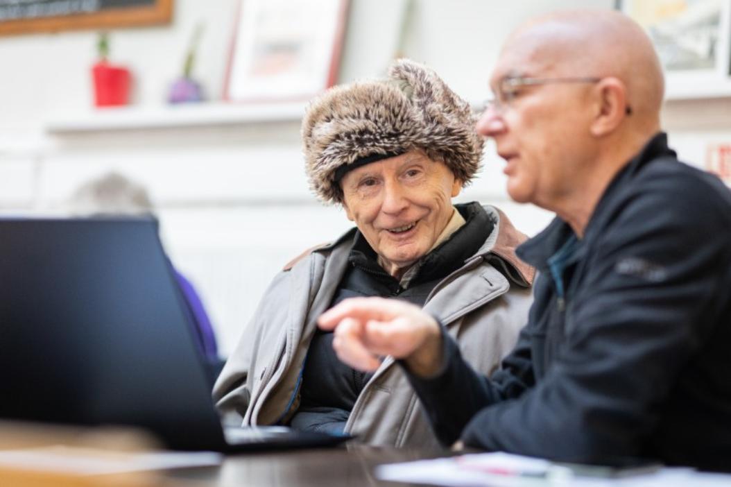 An older gentleman wearing a wooly hat facing toward the camera while sitting in front of a laptop, while another gentleman in the foreground is pointing at the screen and speaking